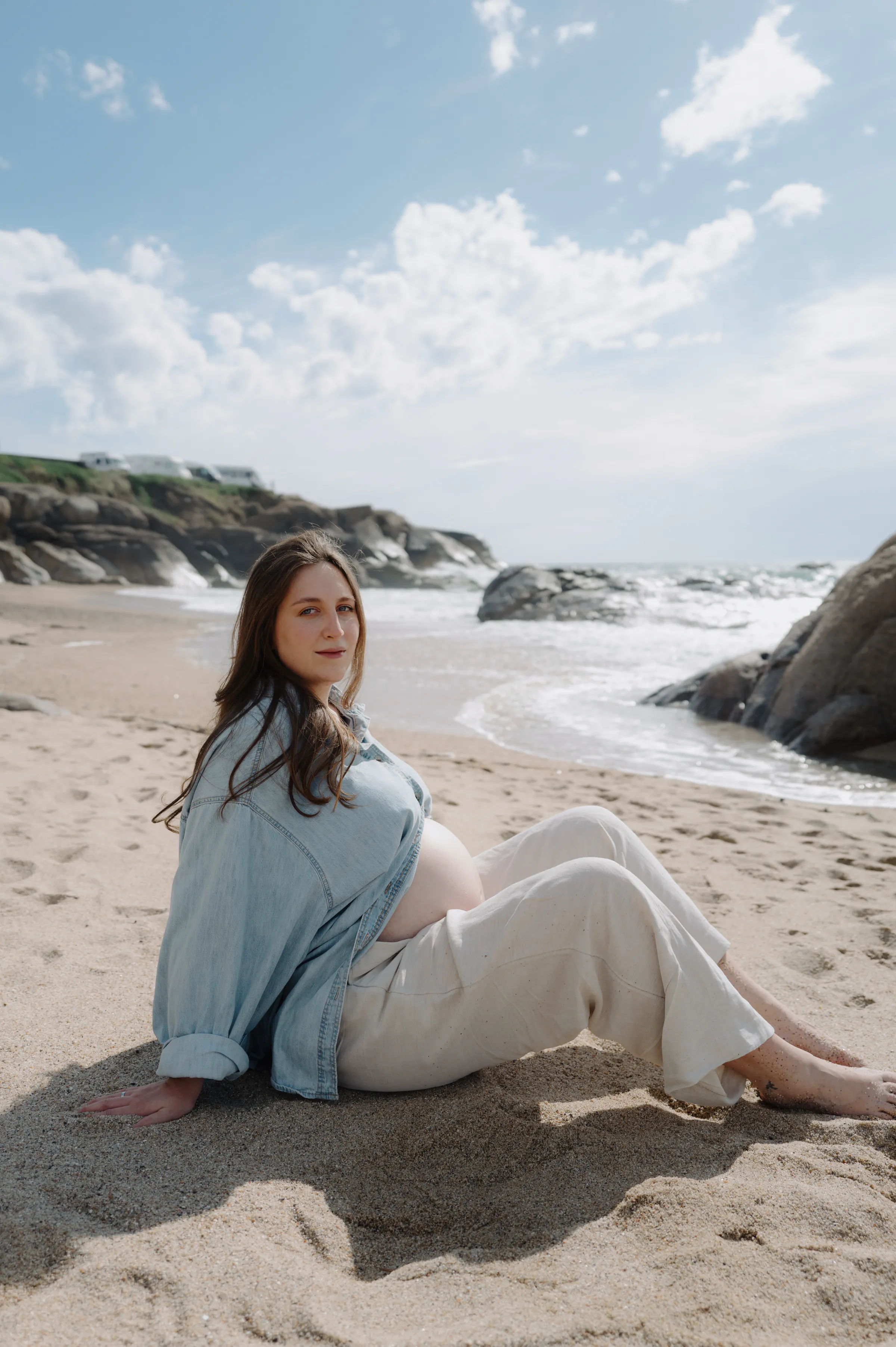 Seaside portrait of an adult seated on sand near rocks and waves in soft daylight.