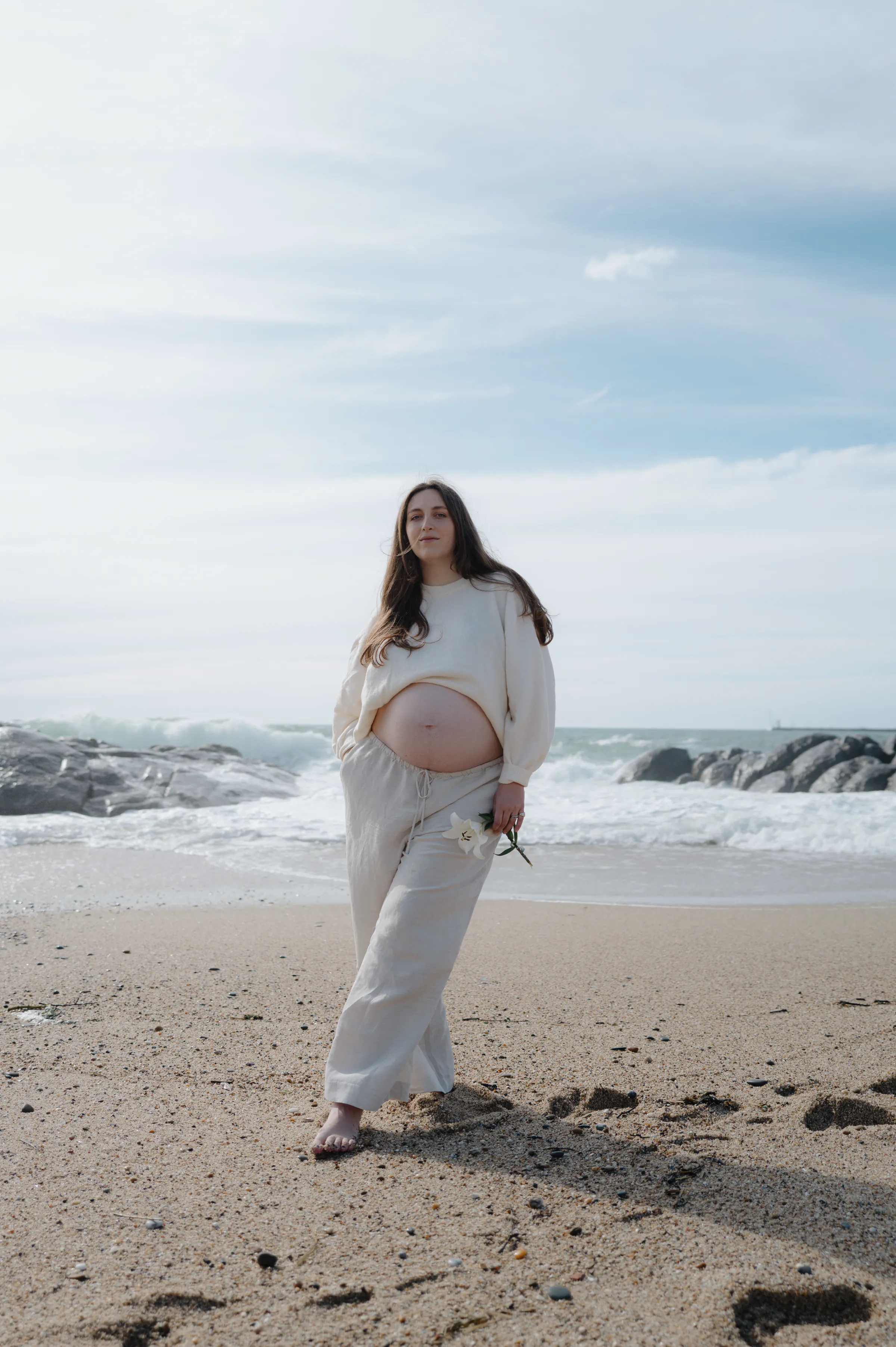 Seaside portrait of a person in white standing barefoot on sand near rocks and waves.