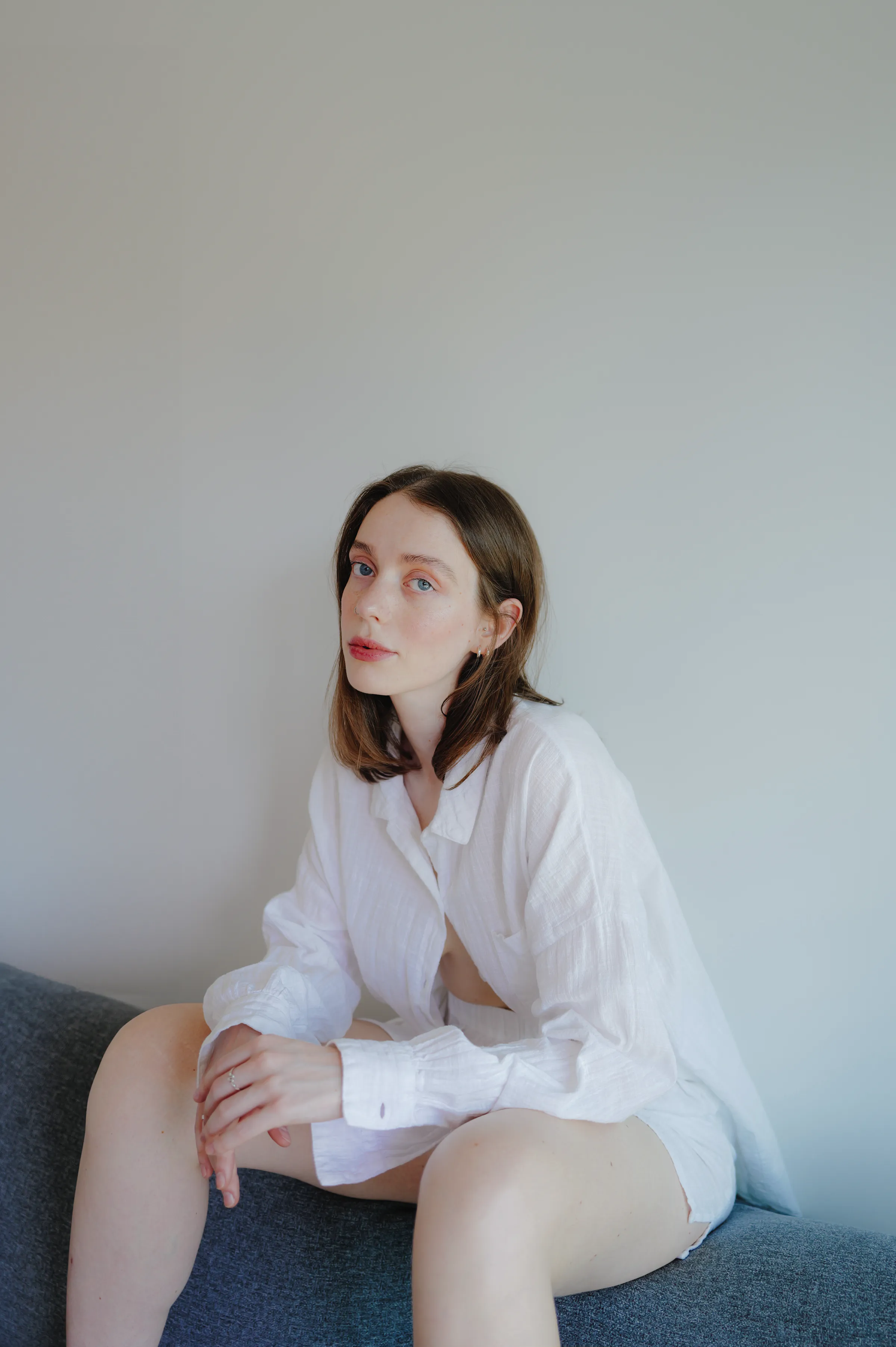 Indoor portrait of a seated person in a white shirt on a gray sofa against a plain wall.