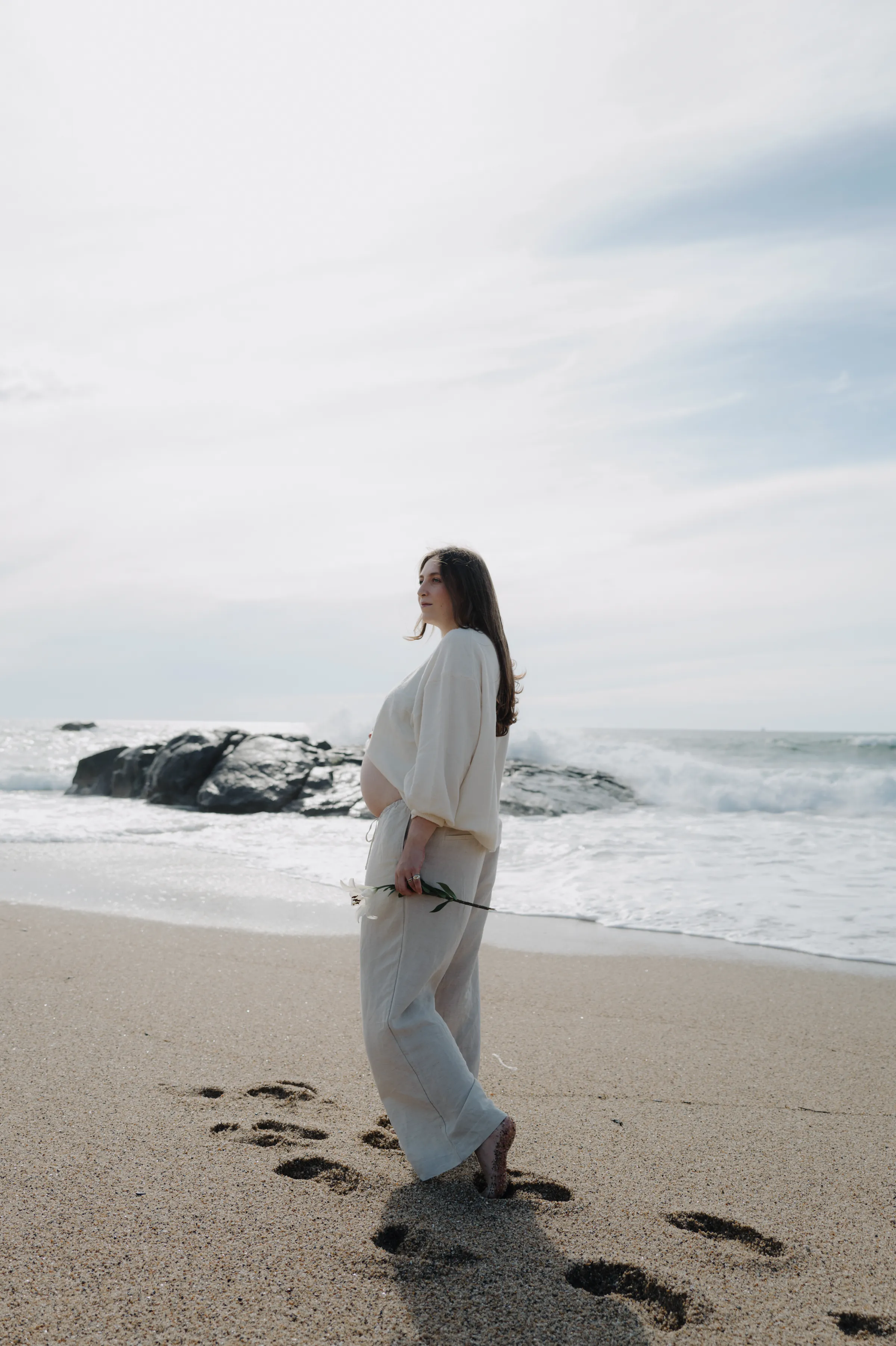 Seaside portrait of a person in white standing by the waves with footprints in the sand.