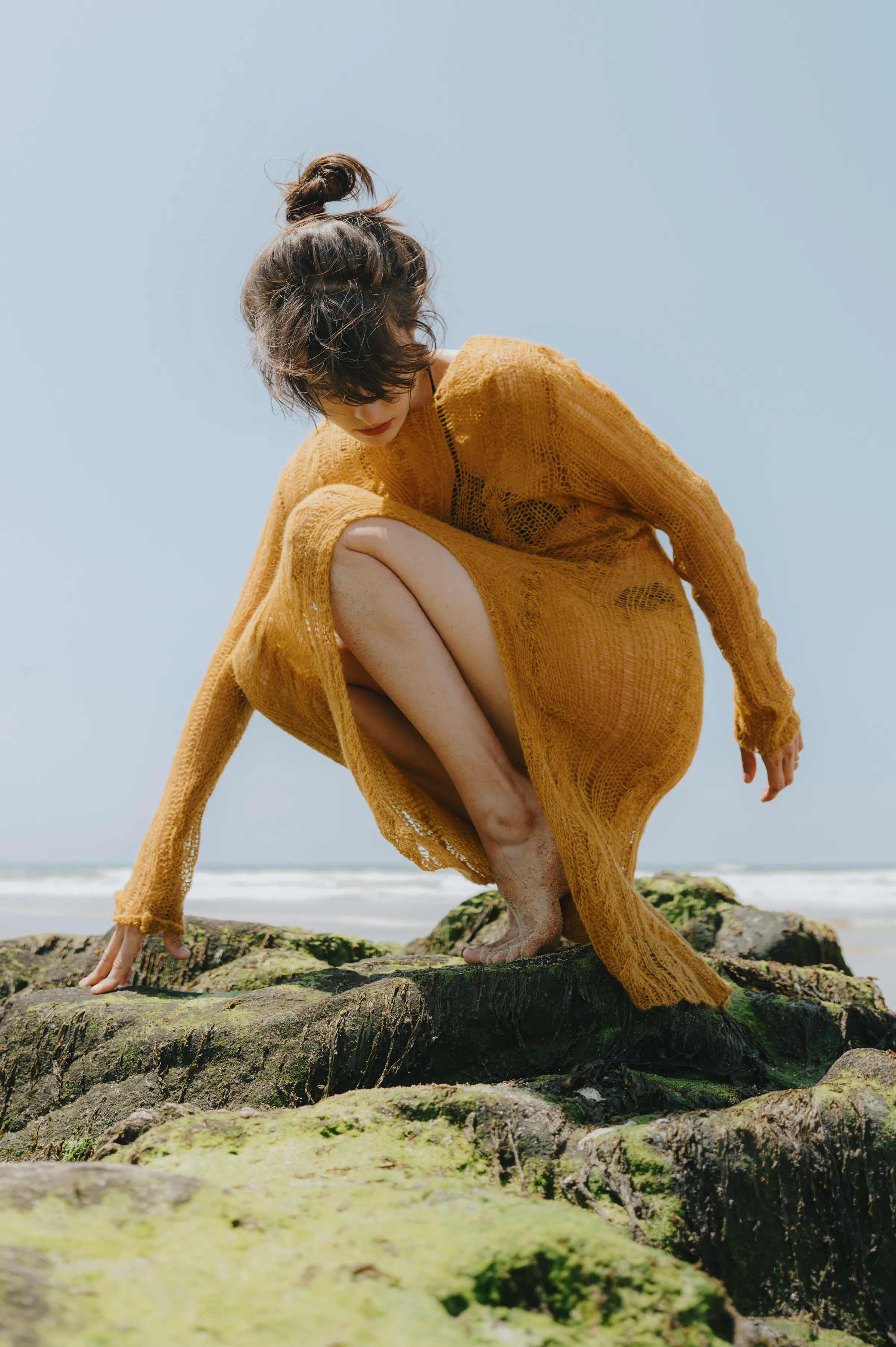 Outdoor seaside portrait of a person in a mustard dress crouching on mossy rocks by the water.
