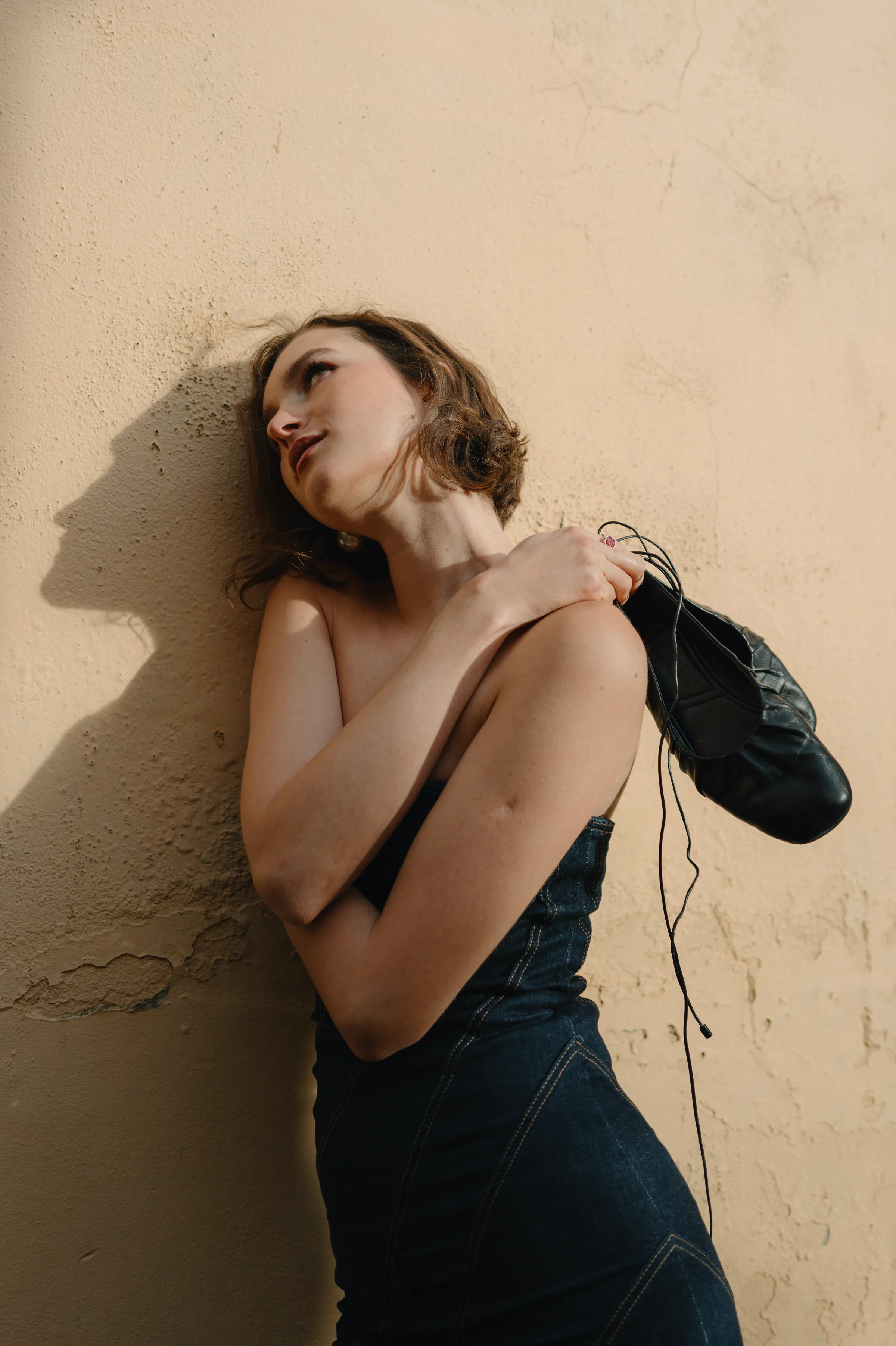 Portrait of an adult leaning against a textured wall, holding black shoes in soft side light.