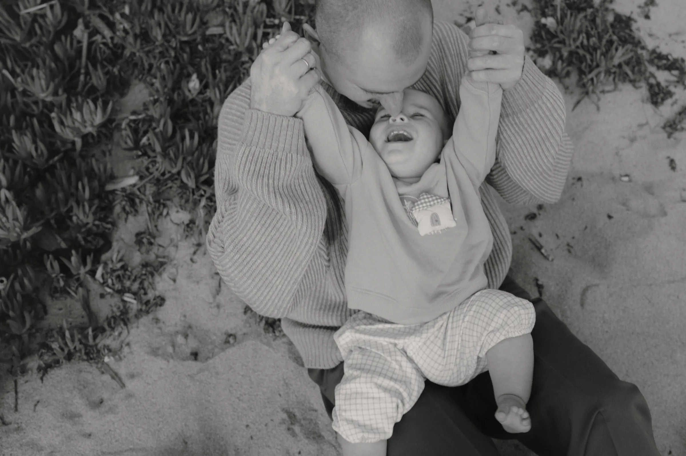 Black-and-white outdoor portrait of an adult holding a baby's hands on sand beside low shrubs.