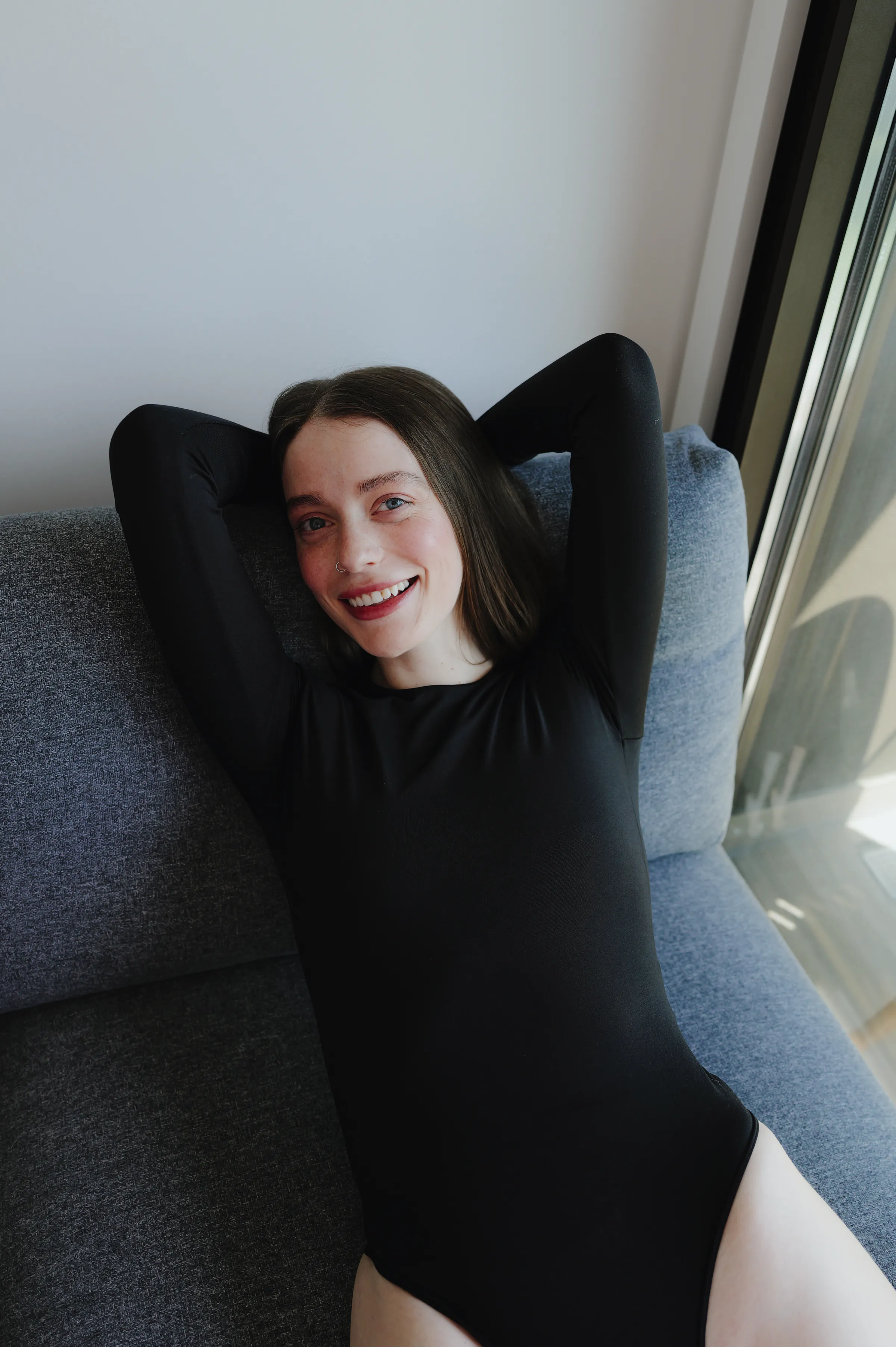 Close portrait of a woman reclining on a gray sofa by a window in soft indoor light.