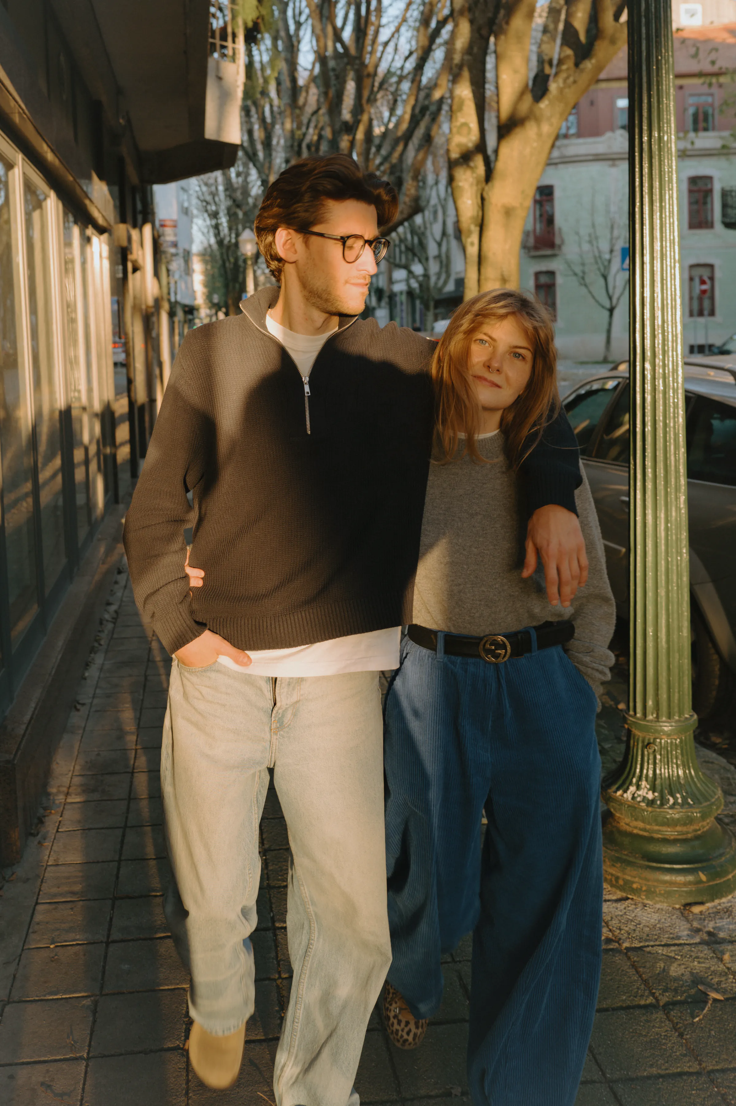 Couple portrait during a city walk, standing close on a sunlit sidewalk beside storefronts and bare trees.