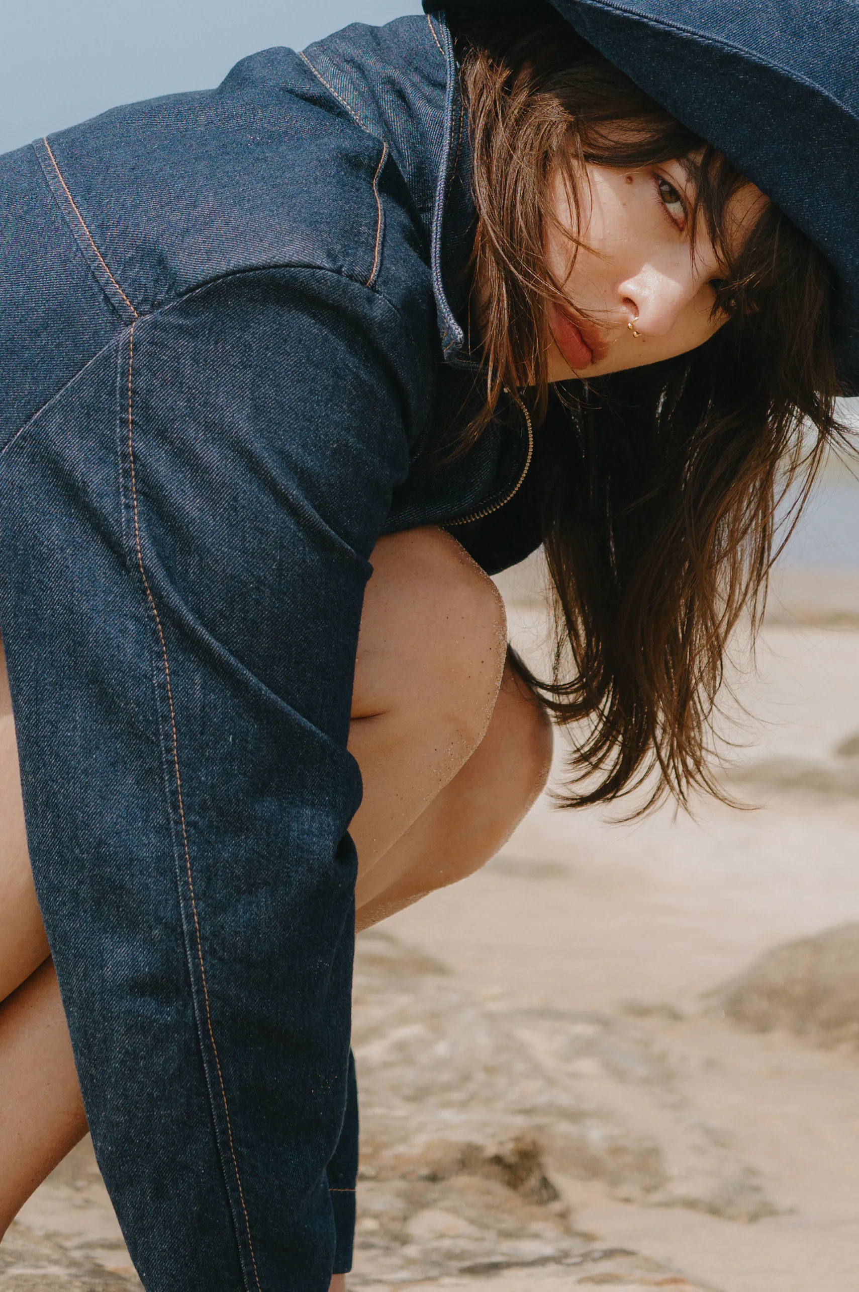 Close portrait of a person crouching on a sandy seaside in a denim hooded jacket, soft light.