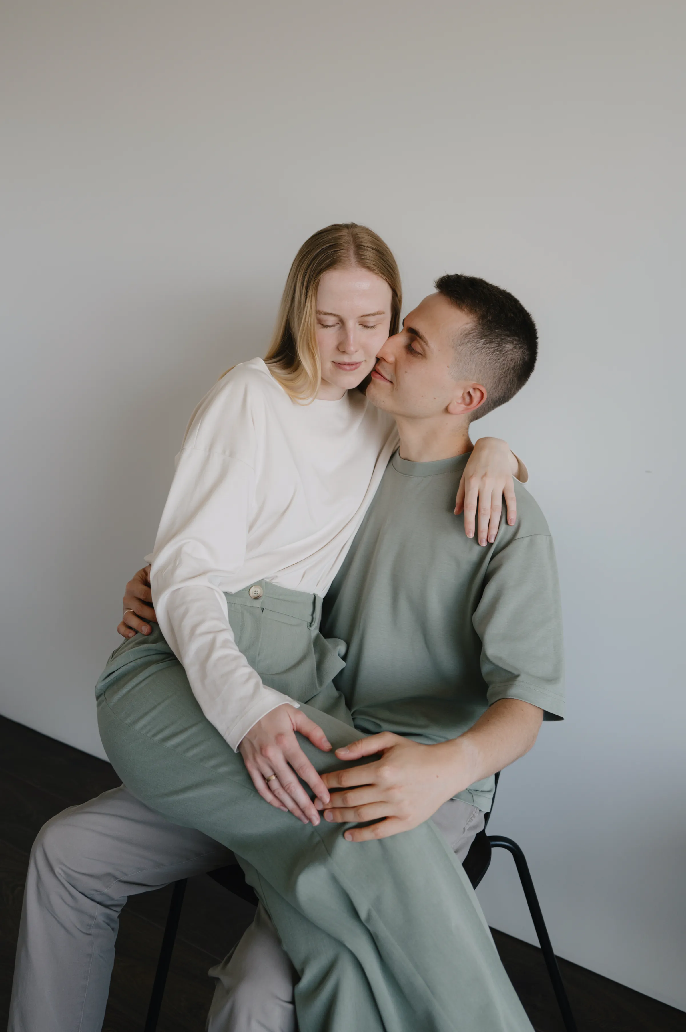 Couple seated closely in a minimalist indoor portrait, hands resting on a rounded belly in soft light.