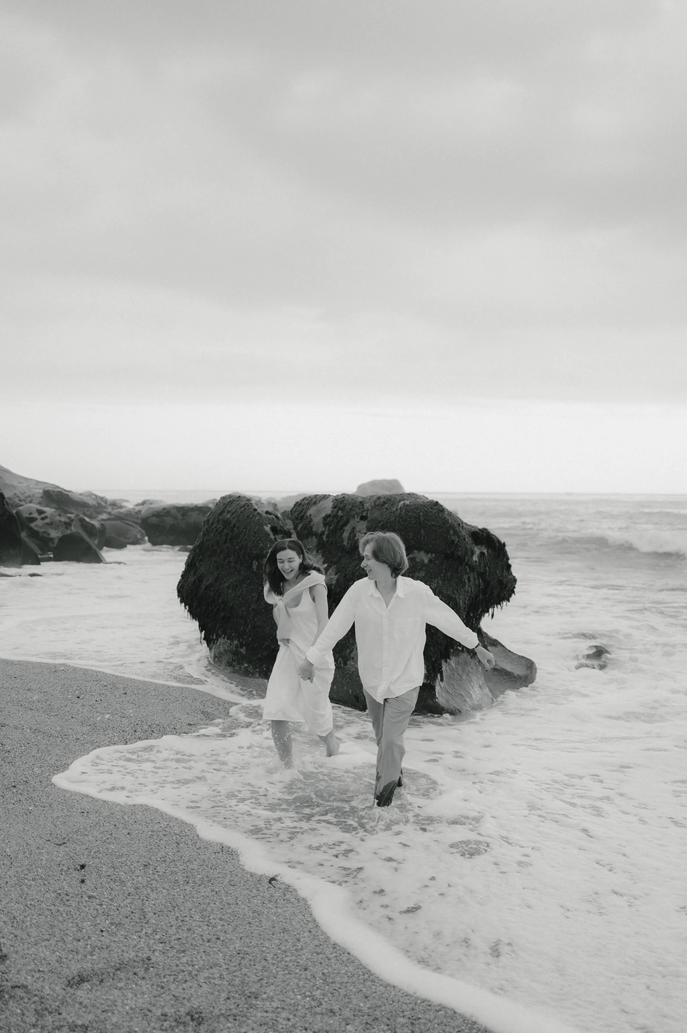 Couple holding hands during a seaside session, walking through shallow surf near dark rocks in soft overcast light.