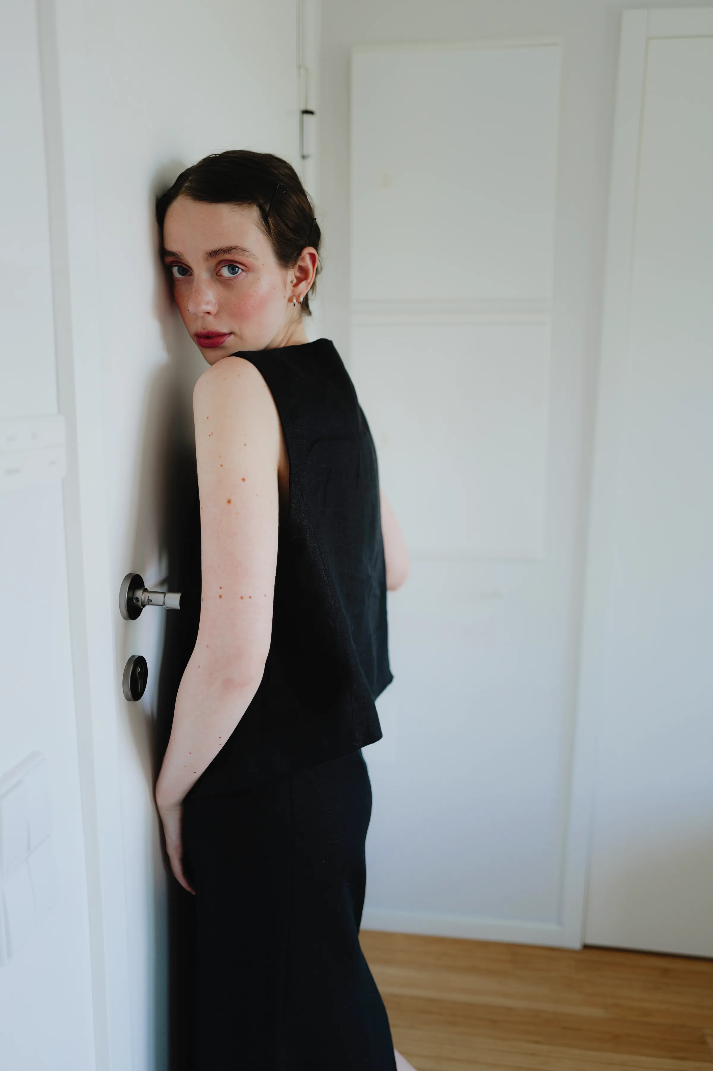 Indoor portrait of an adult in black leaning beside a white door in soft light