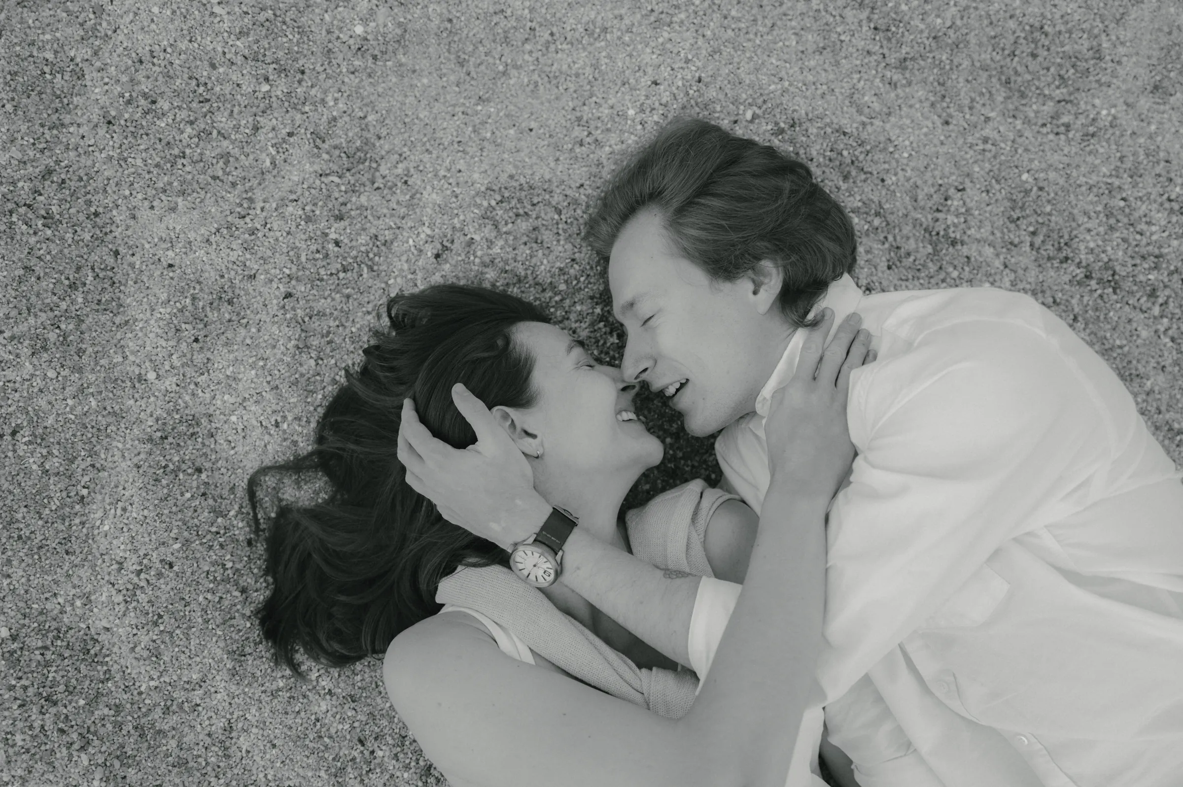 Black-and-white close portrait of a couple lying together on a sandy seaside surface.