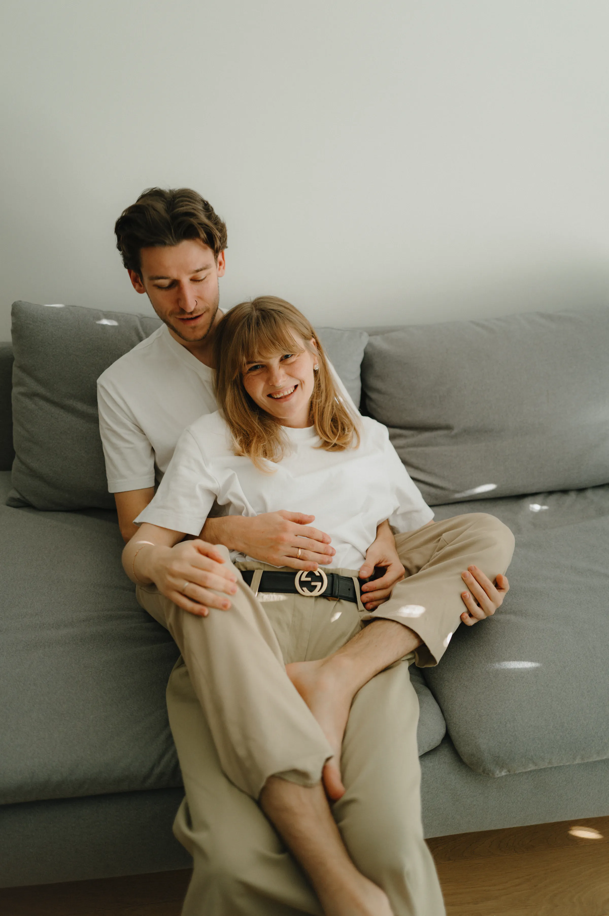 Family home session portrait of an adult and child sitting together on a gray sofa in soft light.