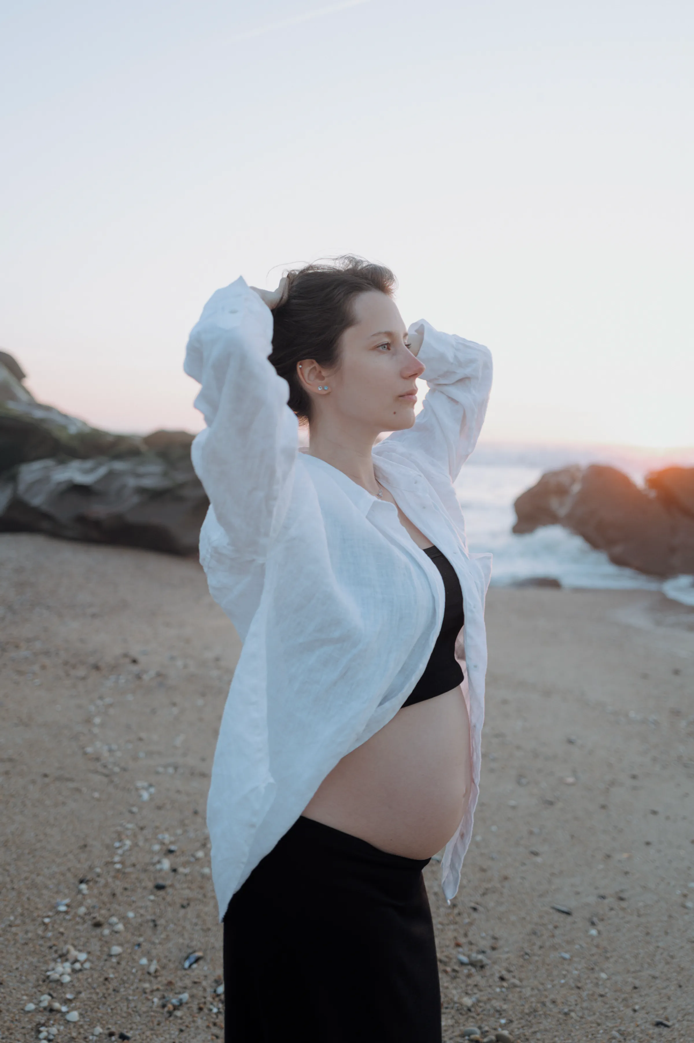 Seaside portrait of an adult in a white cover-up standing on a rocky beach at soft sunset light